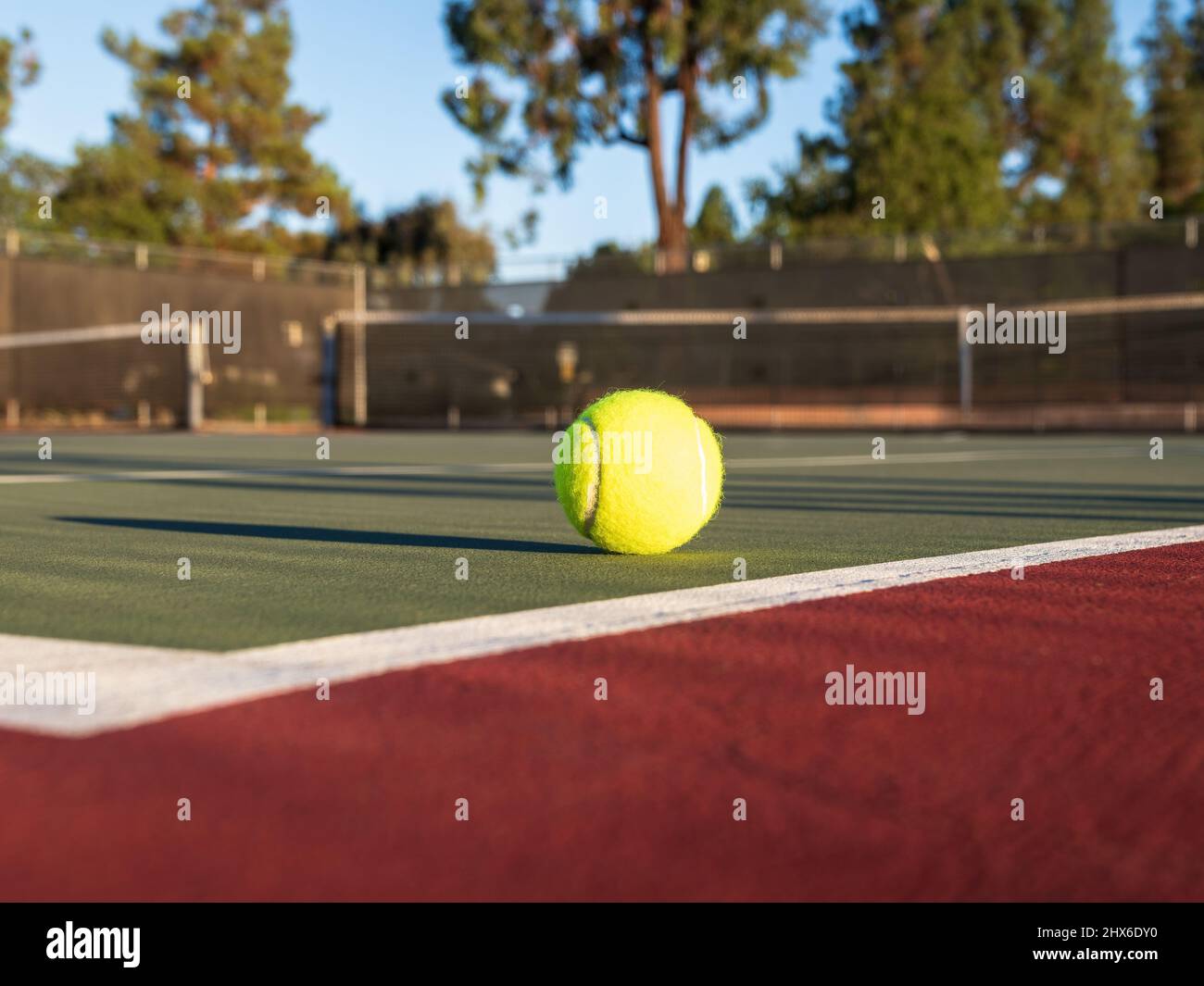 Yellow tennis ball on green court with black net and treelike in background. Red surrounding surface. Stock Photo