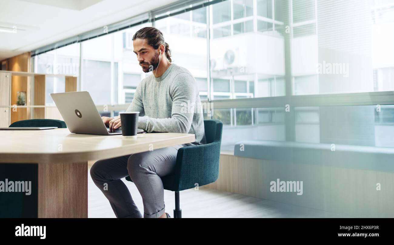 Modern businessman listening to music on his earphones while typing an email. Focused young businessman using a laptop while working remotely in a co- Stock Photo