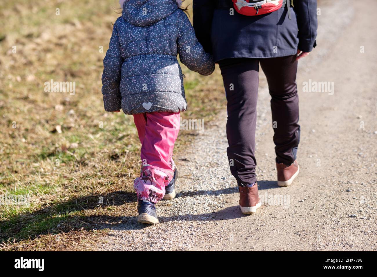 Rear view of low section of a 6 year old child girl in winter clothing walking hand in hand with her mother on a sunny gravel road in Germany in Febru Stock Photo