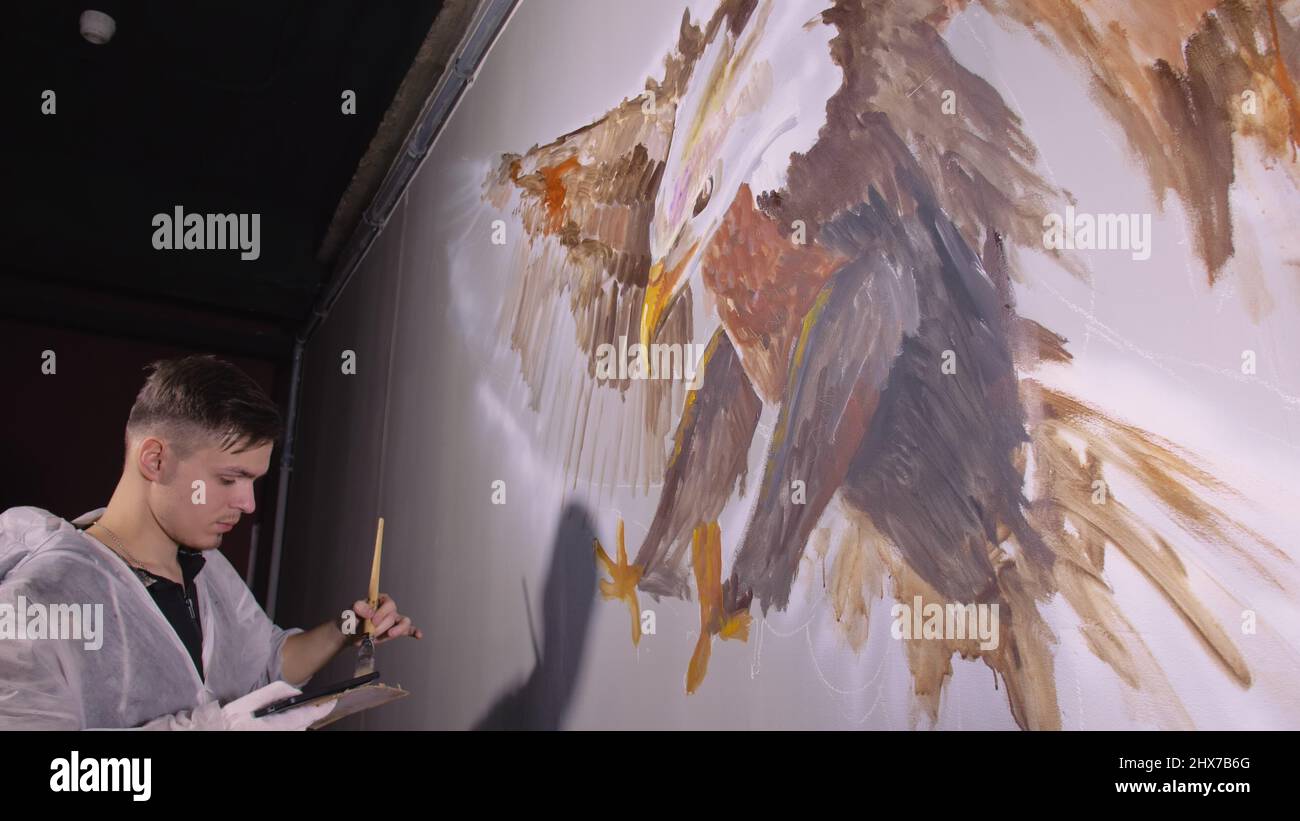 Artist designer draws an eagle on wall. Craftsman decorator paints picture with acrylic oil color looking at sketch in phone. Painter painter dressed Stock Photo