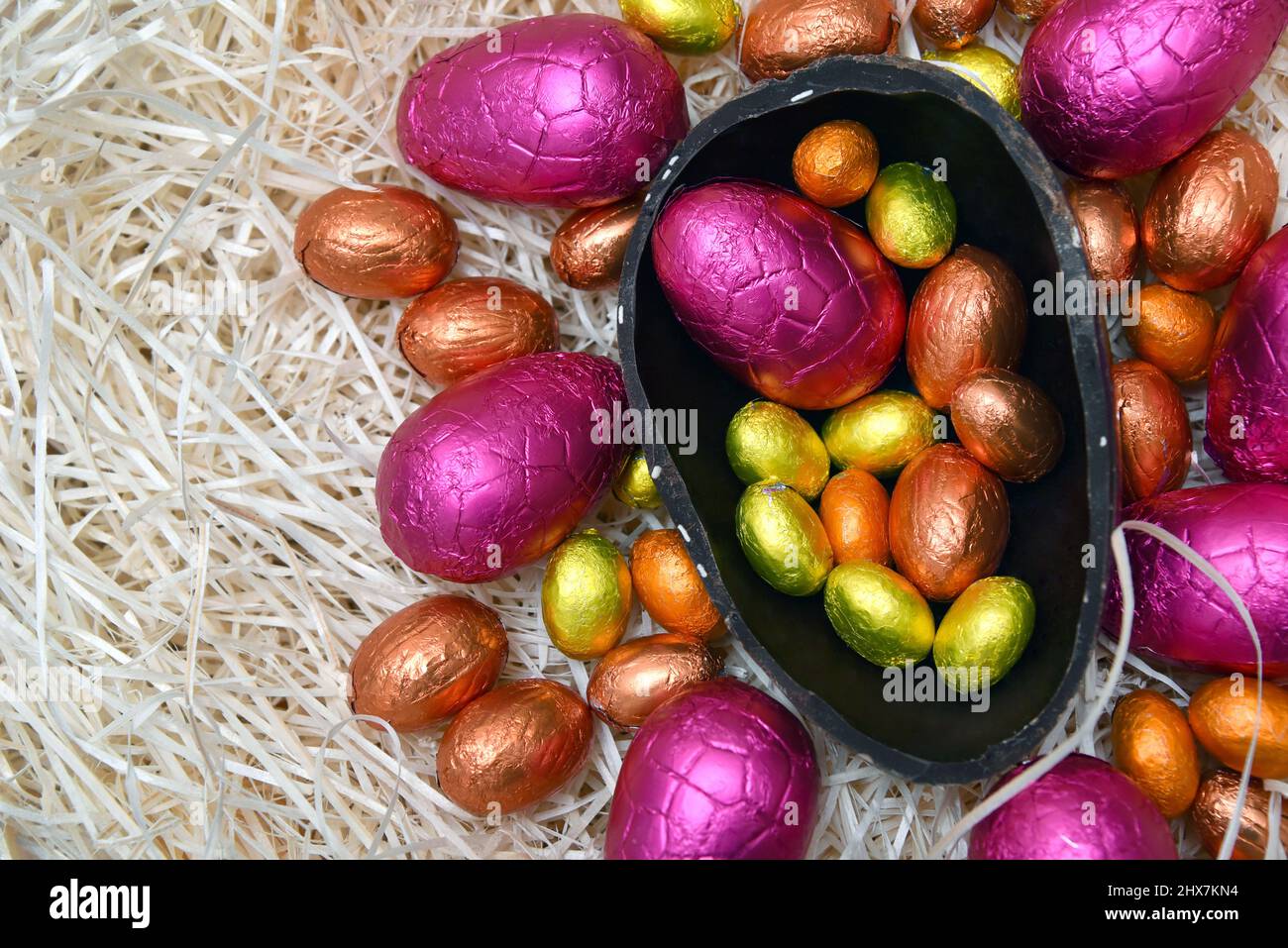Pile of colorful foil wrapped chocolate easter eggs in pink, red, silver and gold with two halves of a large brown dark chocolate egg. Stock Photo