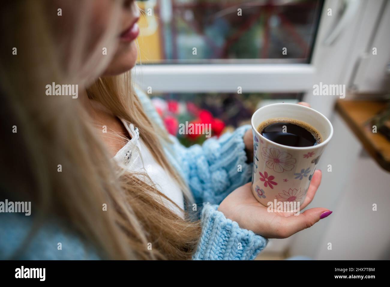 A woman sits at home holding a mug of hot coffee in her hands. Stock Photo