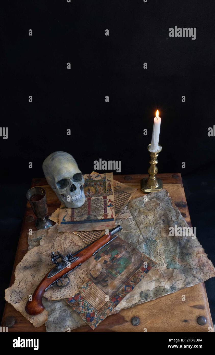 Pirate table, candle, skull, maps old pages goblet and flintlock pistol spread on wooden table. Stock Photo