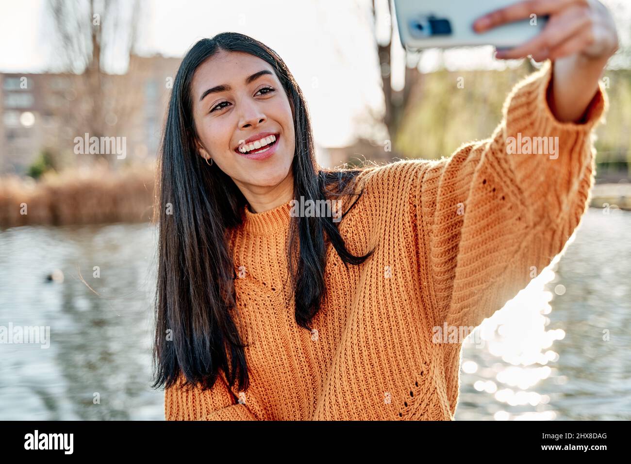 Photo of carefree long haired young latin woman takes selfie portrait on mobile phone, wears yellow colorful jumper, uses technology during leisure ti Stock Photo