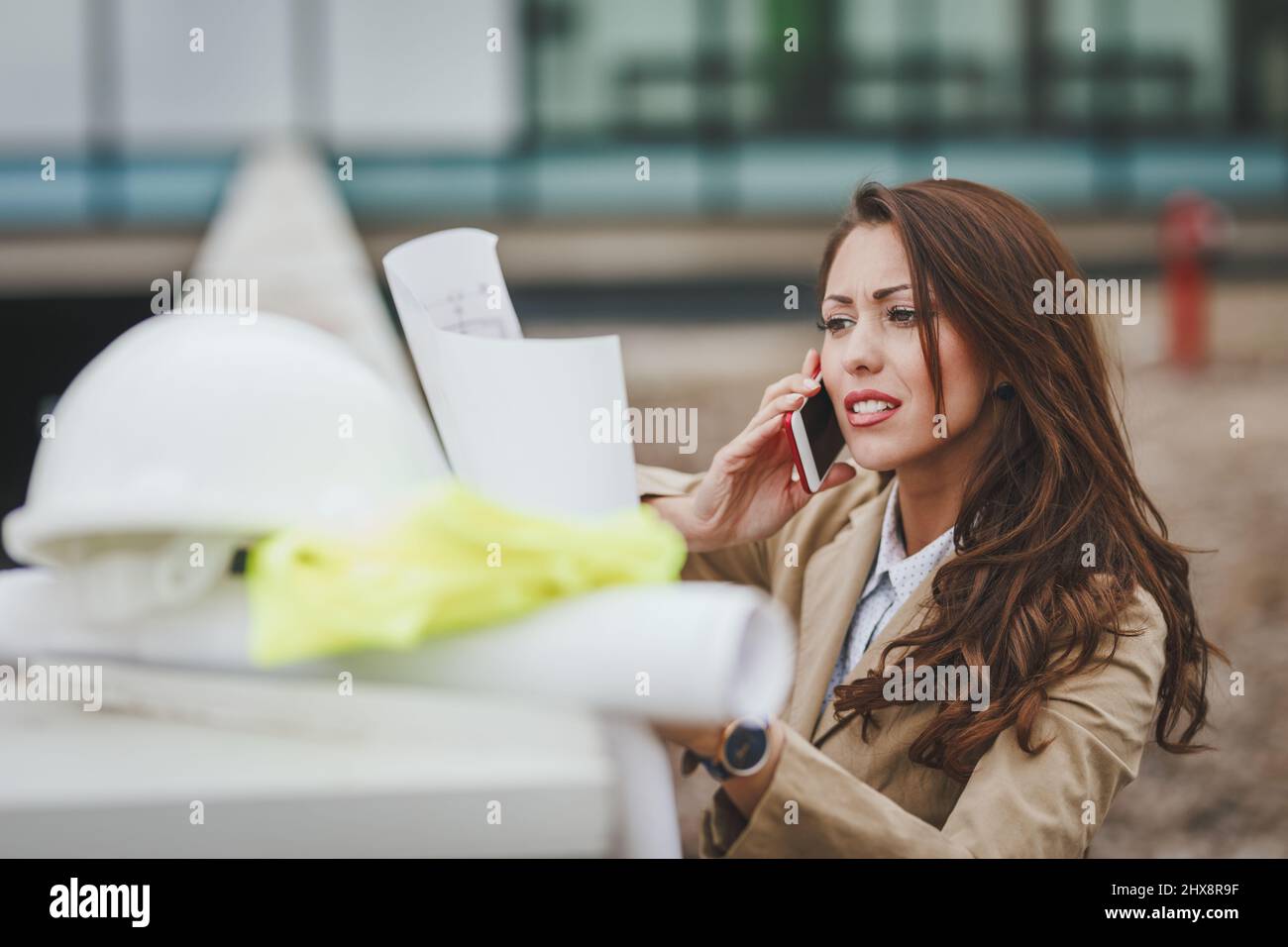 Worriedly successful female engineer with white helmet beside her talking on a smartphone, checked new project and analyzing blueprints in front of co Stock Photo
