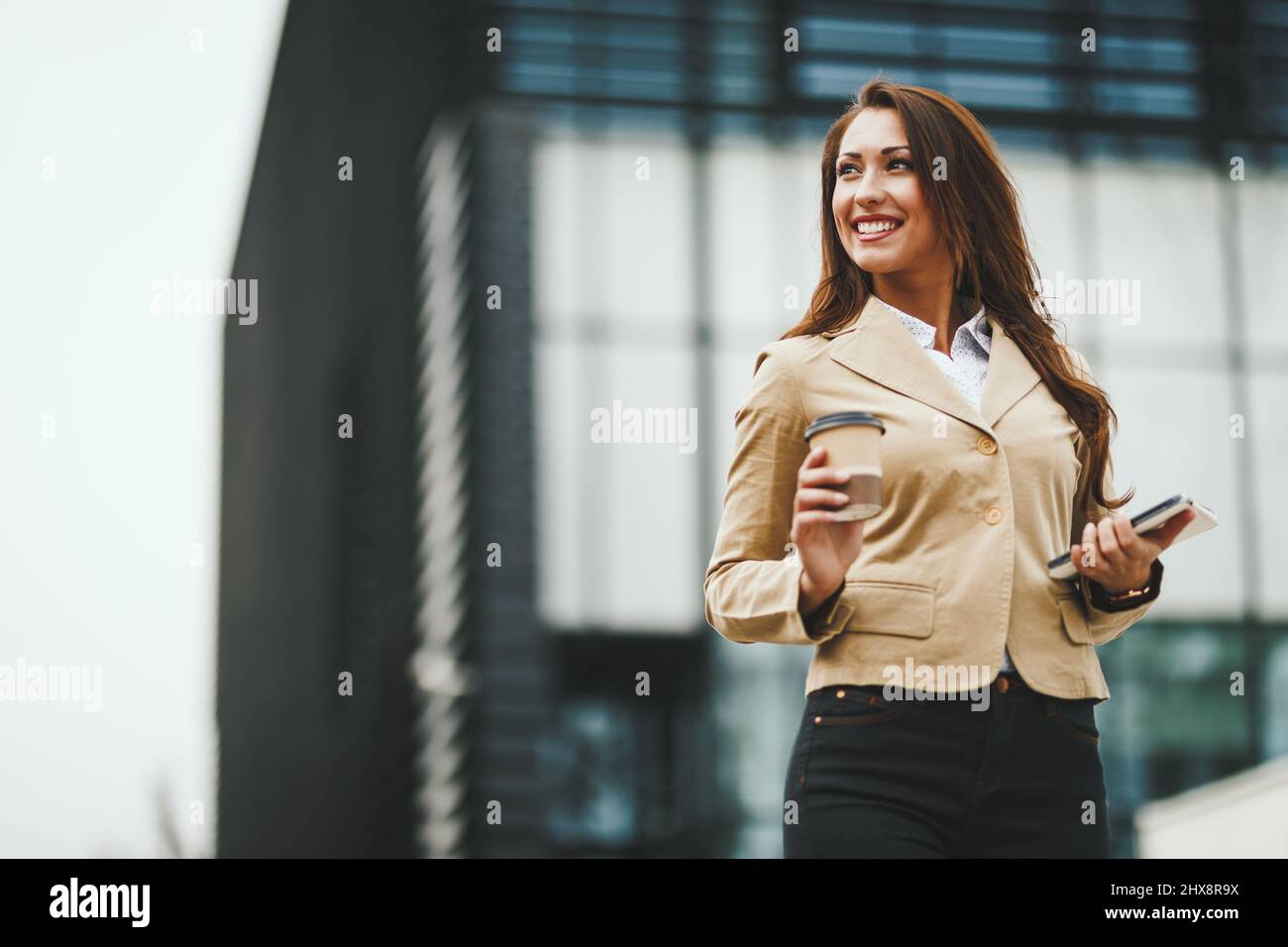 Successful young businesswoman with a cup of coffee to go and digital tablet in her hands standing in front of office district. Stock Photo
