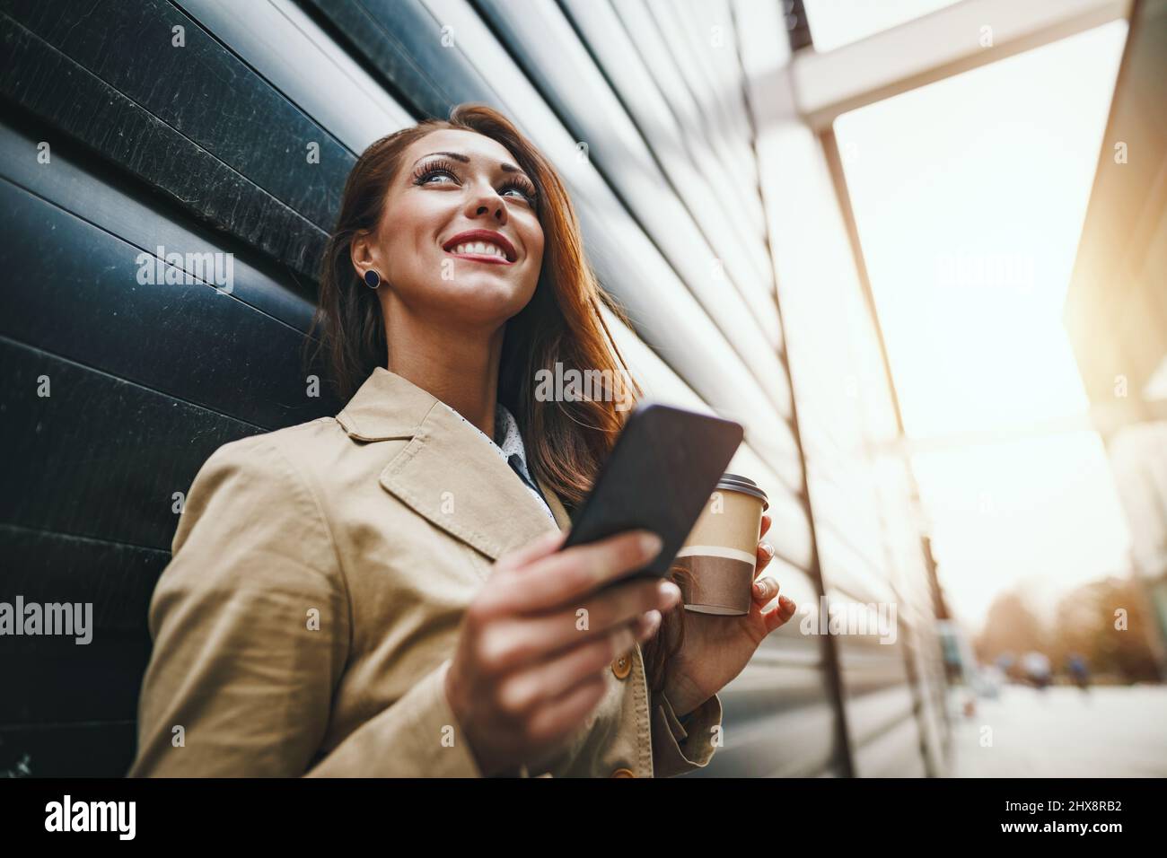 Beautiful young woman with a disposable coffee cup, drinking coffee and using smartphone in the office district. Stock Photo