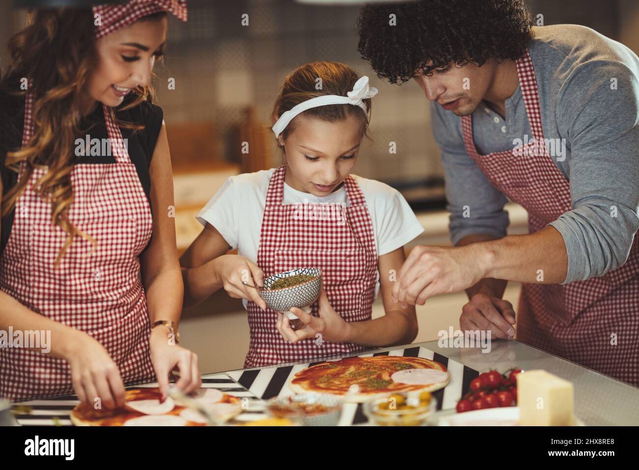 Happy parents and their daughter are preparing meal together in the kitchen while little girl is putting ketchup on the pizza dough. Stock Photo