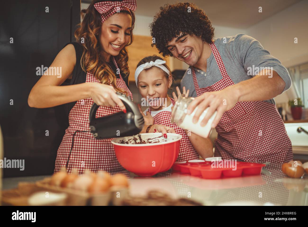 Happy parents and their daughter are preparing cookies together in the kitchen. Little girl helps to her parents mixing dough with mixer. Stock Photo