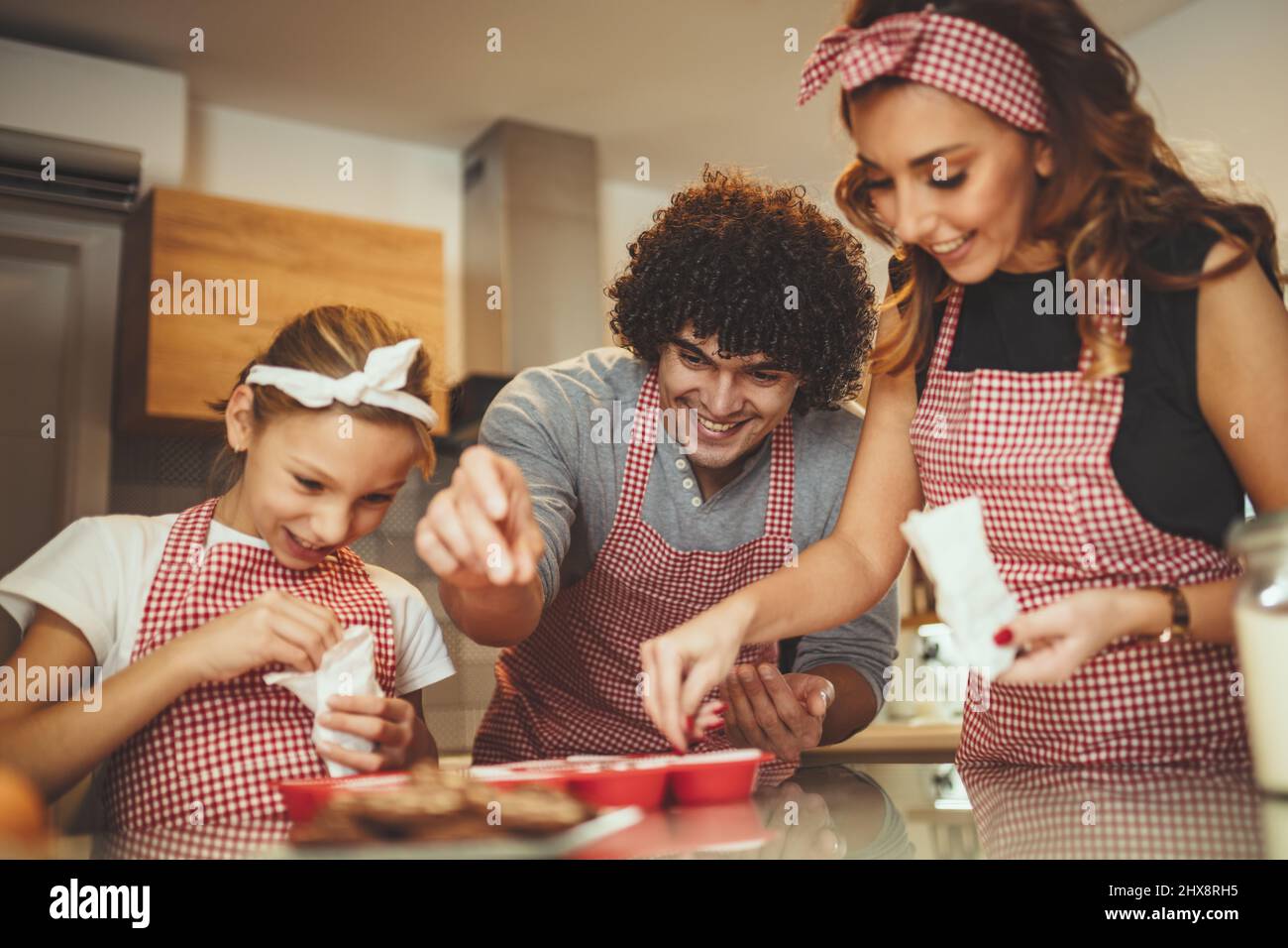 Happy parents and their daughter are preparing cookies together in the kitchen. Little girl helps to her parents to put crumbs of chocolate on the coo Stock Photo