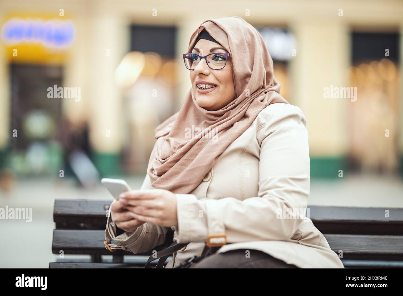 Middle aged Muslim woman wearing hijab with a happy face is sitting on the bench in urban environment, tipping messages on her smartphone. Stock Photo