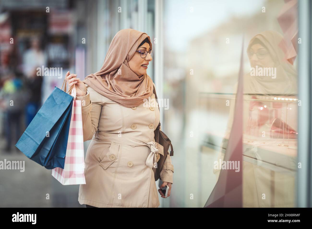 Middle aged Muslim woman wearing hijab with a happy face is walking in urban environment, going to the shopping holding paper colored bags. Stock Photo