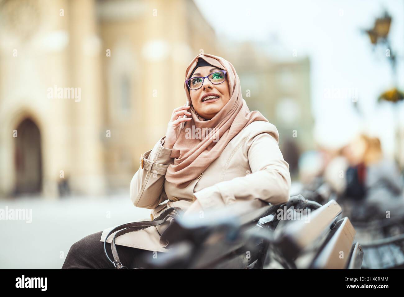 Middle aged Muslim woman wearing hijab with a happy face is sitting on the bench in urban environment, talkings on her smartphone. Stock Photo