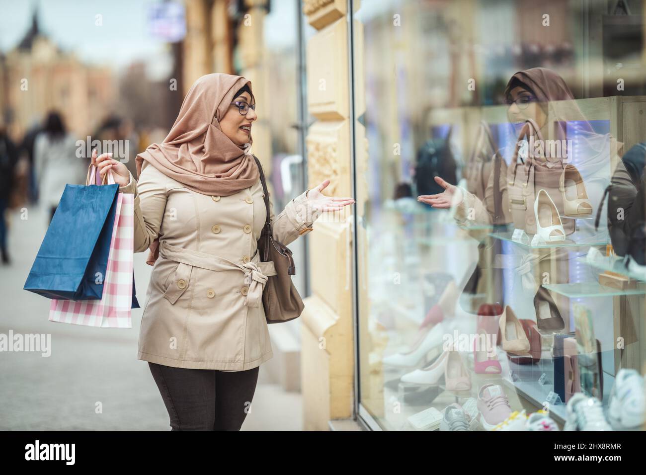 Middle aged Muslim woman wearing hijab with a happy face is walking in urban environment, going to the shopping holding paper colored bags. Stock Photo