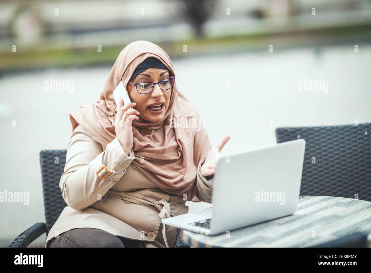 Middle aged worried Muslim woman wearing hijab is sitting on the cafe in urban environment, talking by smartphone and working on laptop. Stock Photo