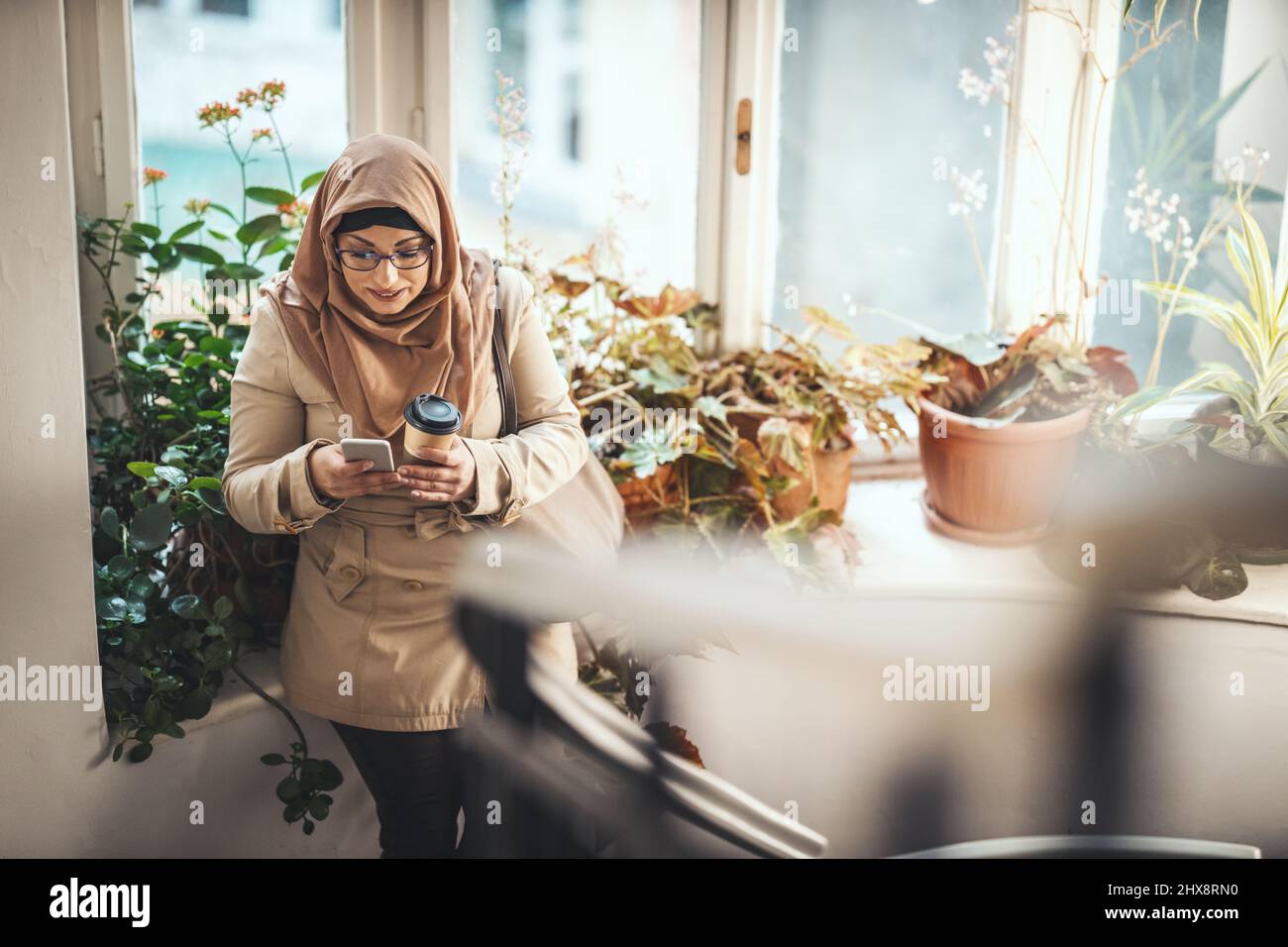Middle aged Muslim woman wearing hijab with a happy face is standing by the window tipping messages on her smartphone. Stock Photo
