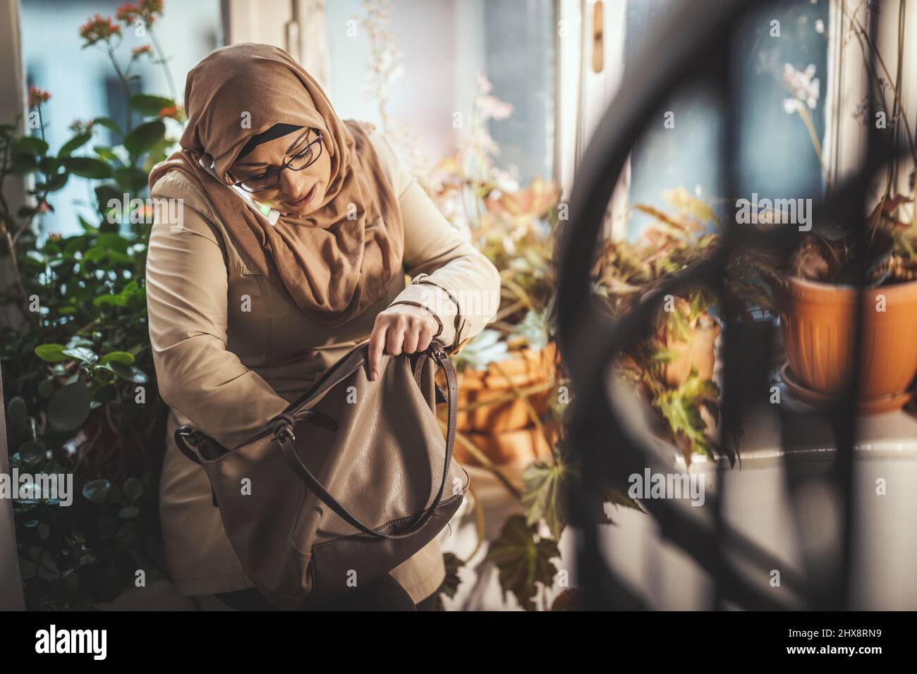 Middle aged Muslim woman wearing hijab with a happy face is standing by the window looking for something in her purse and taking on her smartphone. Stock Photo