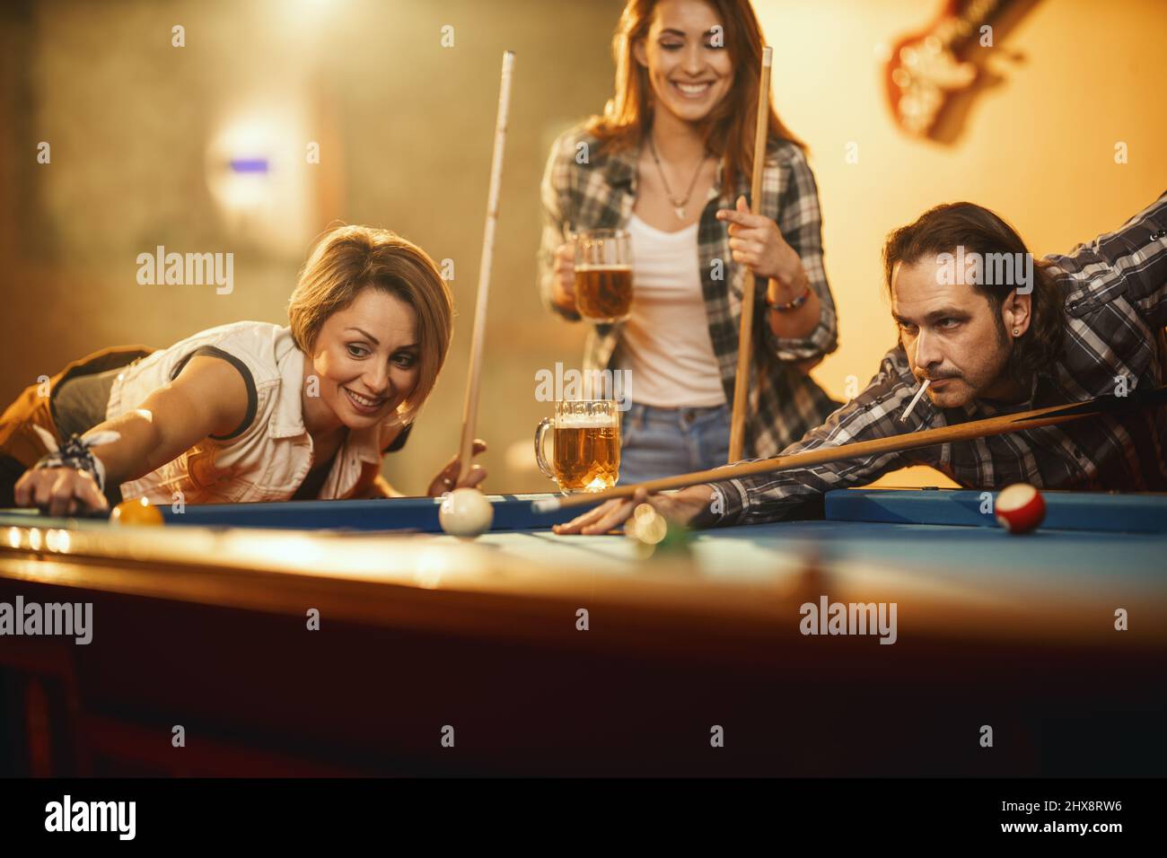 Young smiling cheerful friends are playing billiards in bar and drinking beer after work.  They are involved in recreational activity. Stock Photo