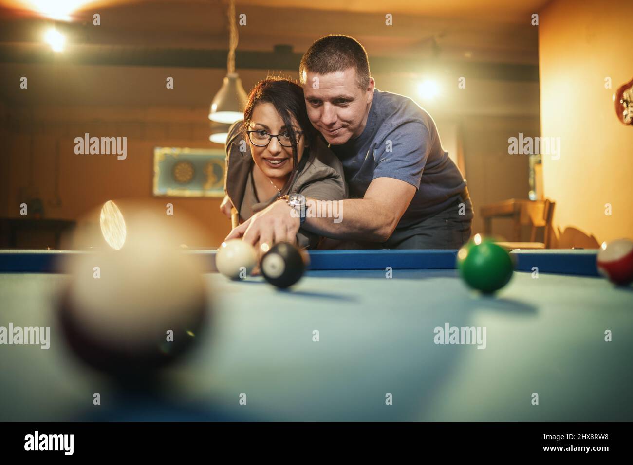 Young smiling cheerful couple is playing billiards in bar after work. They are having fun and involved in recreational activity. Stock Photo