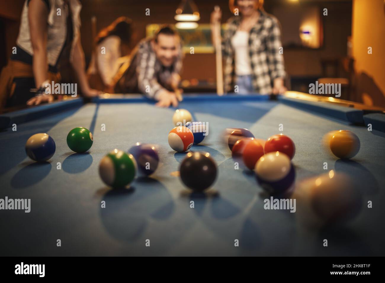 Young smiling cheerful friends are playing billiards in bar after work. They are involved in recreational activity. Selective focus, focus on billiard Stock Photo