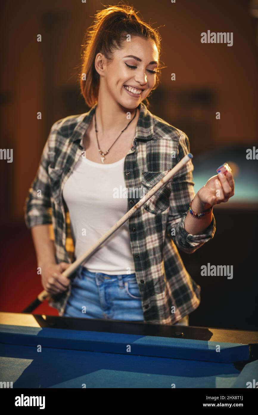 Smiling young woman apply chalk to the end of their cue sticks in billiard club. Stock Photo