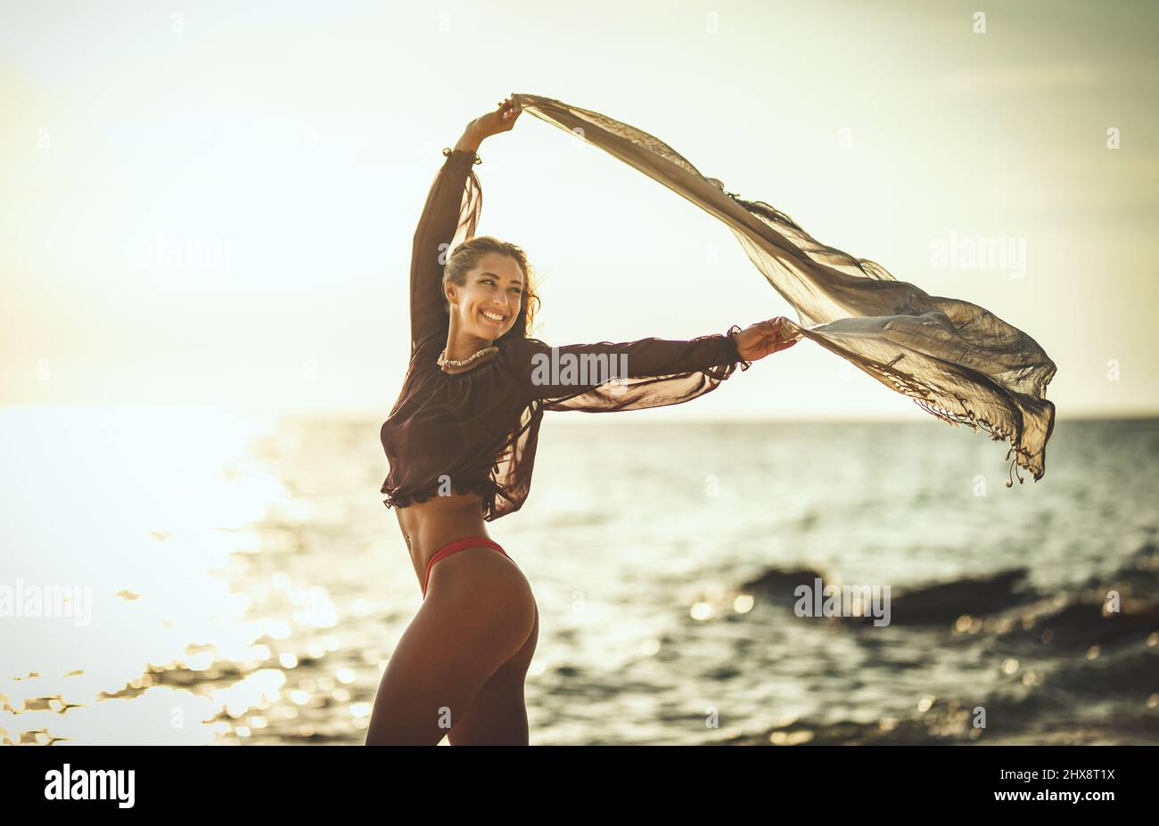 A beautiful young woman is having fun and relaxing on the beach at the sunset. She is posing and looking away with transparent scarf in her hands. Stock Photo