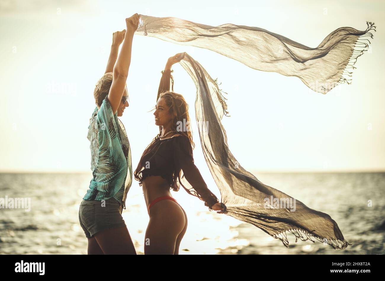 Beautiful two young women are having fun and relaxing on the beach at the sunset. They are posing and looking each other with transparent scarfs in th Stock Photo