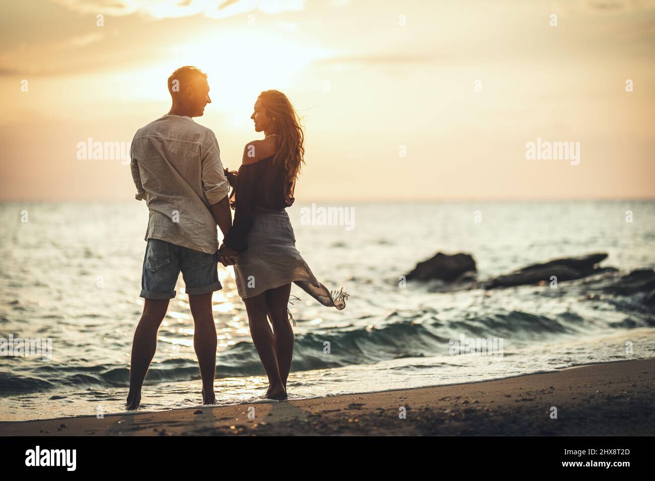 A loving couple is having fun and hugging on the empty sandy sea beach at sunset.They are looking each other and happily smiling. Stock Photo