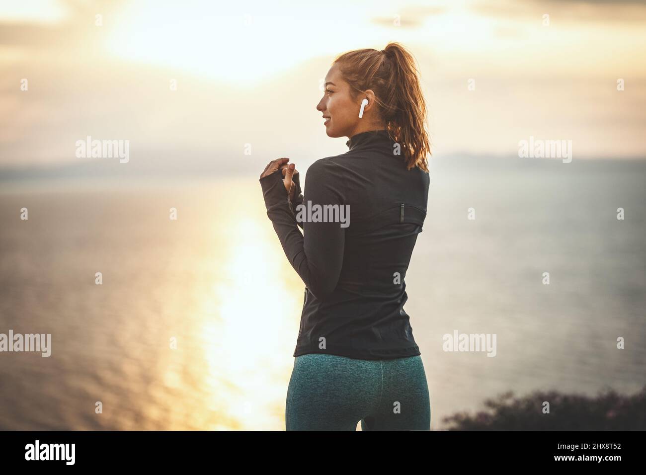 A beautiful young woman is ready to do stretching exercise by the sea in sunrise hearing music by headphones. Stock Photo
