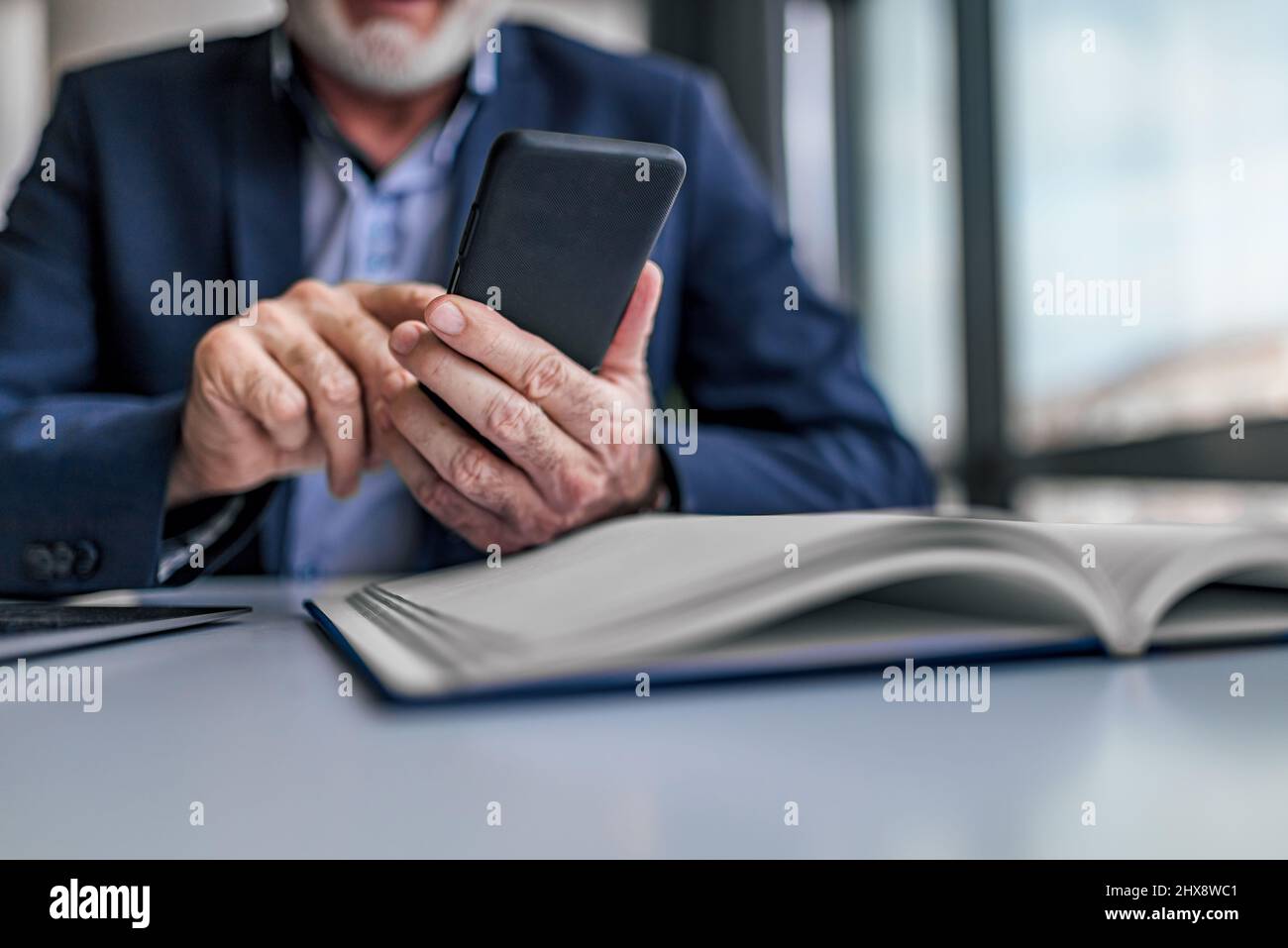Faceless senior man holding smartphone checking messages and emails while sitting at modern bright office. Focus on hand and phone. Stock Photo