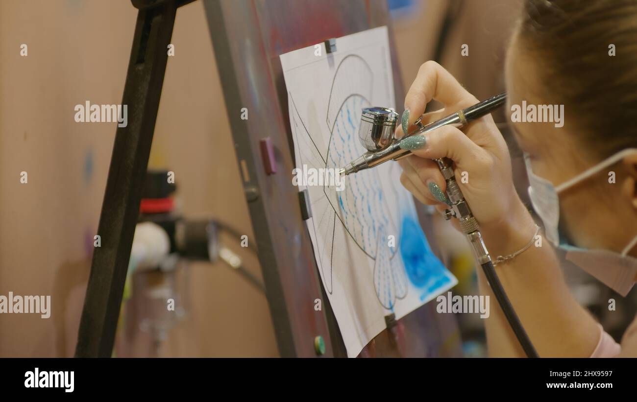 Woman artist learns to paint with airbrush with acrylic dye, paper and easel. Indoors. Concept modern art, airbrushing, aerography, draw spraying pict Stock Photo