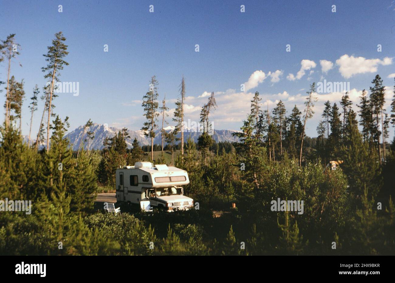 An RV camper parked in a wooded area with mountains in the background ...