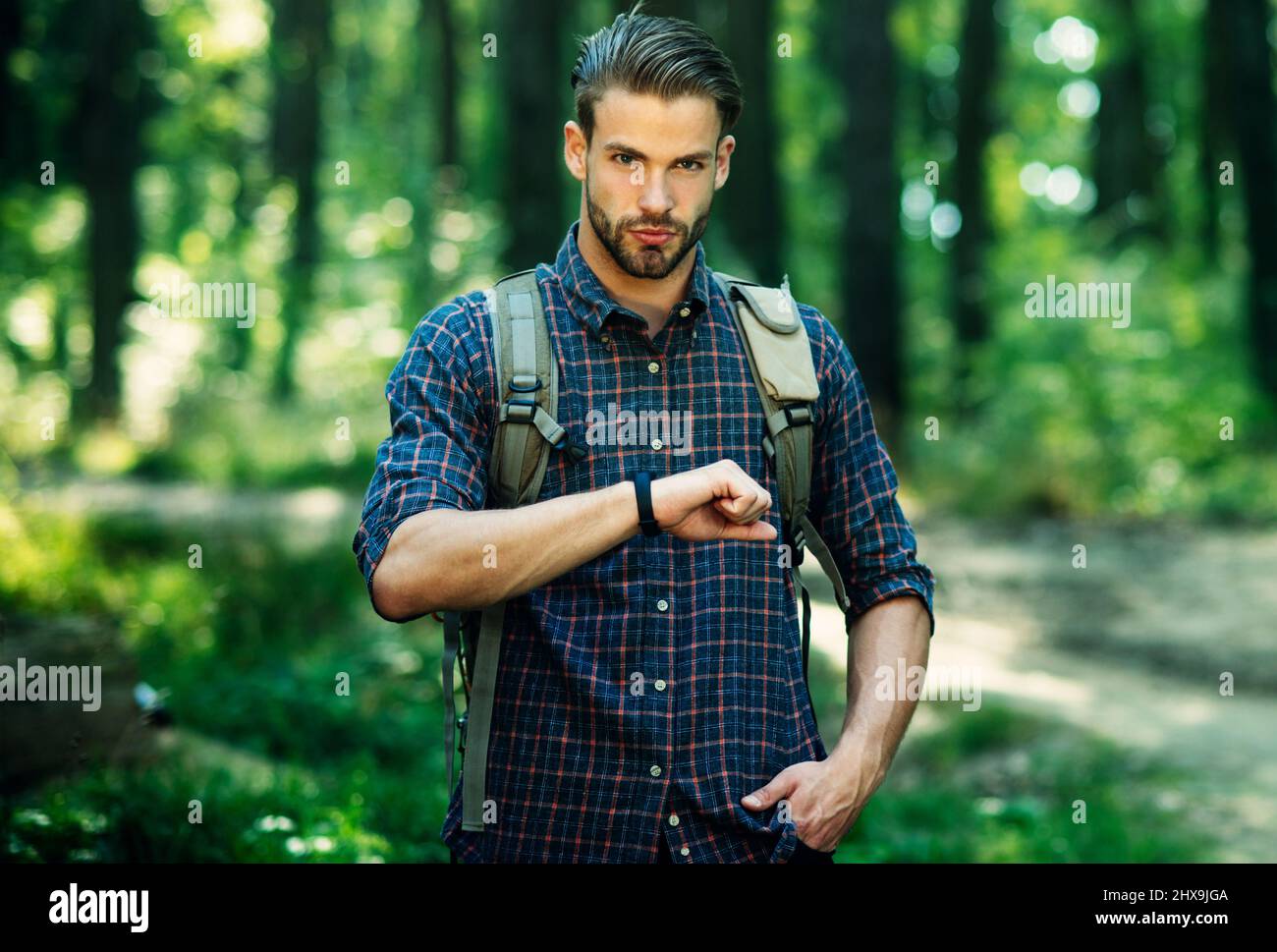 Handsome bearded man with backpack in checkered shirt looks at clock walk in forest. Tourism, travel Stock Photo