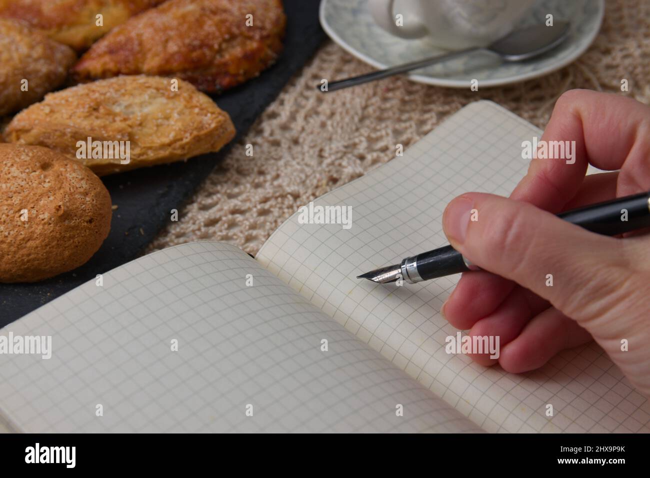 Someone is holding a pen over a notebook with an empty page next to some sweets and a cup of coffee Stock Photo