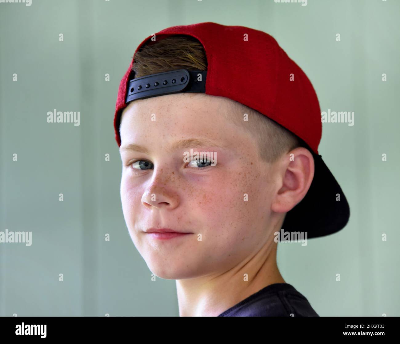 Young boy looks at camera with his baseball cap turned backwards.  He has a small smile on his face.  Freckles sprinkle across nose and cheeks.  Cap i Stock Photo