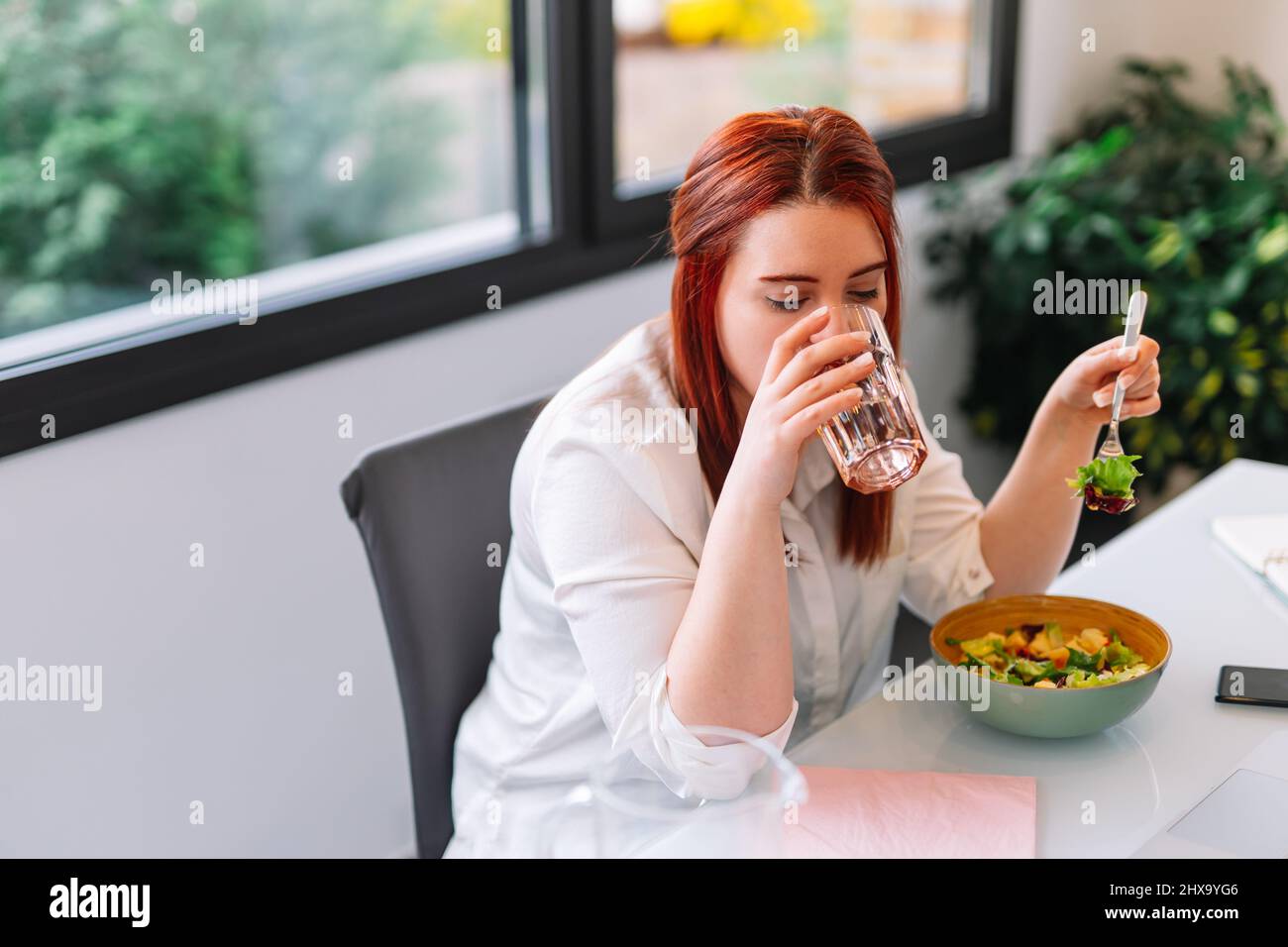 Young woman drinking a glass of water while eating a salad at work