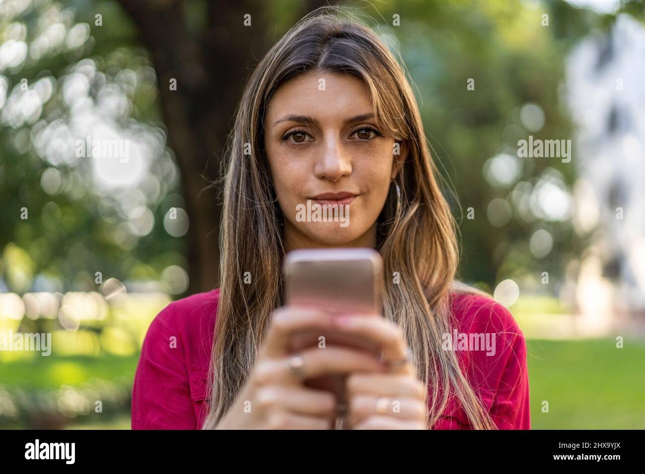 A woman using her smartphone outdoors. She is looking at the camera. Stock Photo