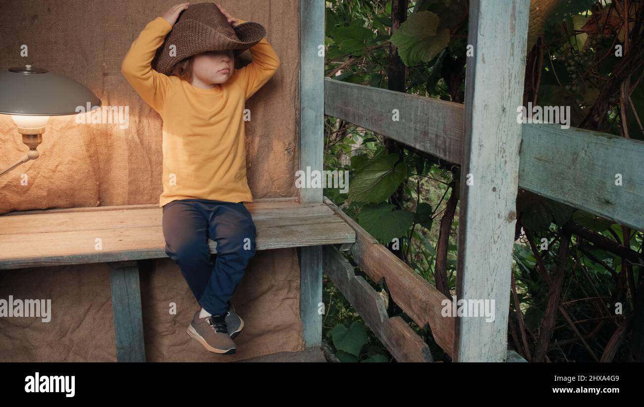 Small, funny child in big cowboy hat is sitting on wooden porch of country house Stock Photo