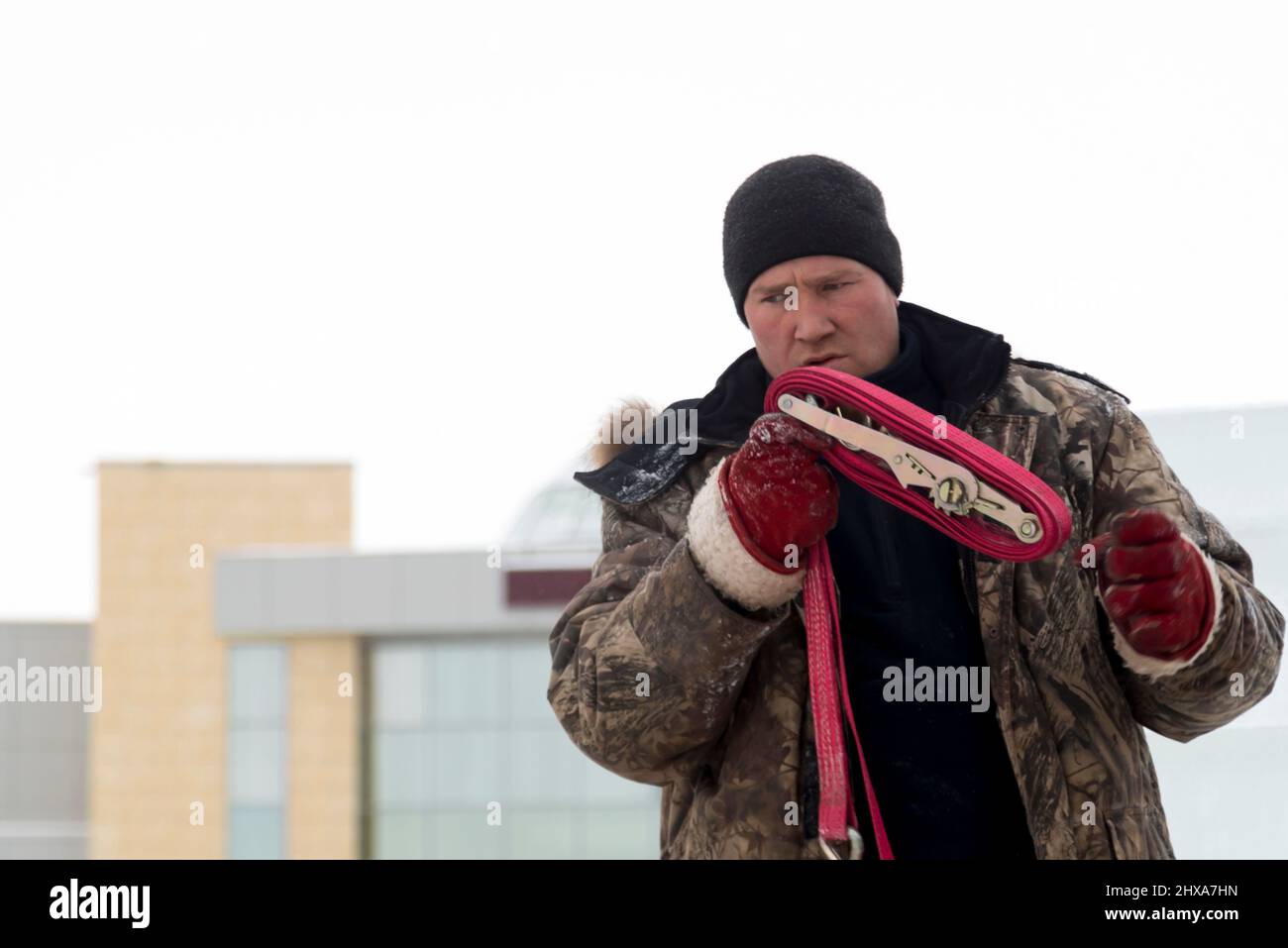 Portrait of a worker in a winter jacket and a black knitted hat with a tape sling in his hands Stock Photo