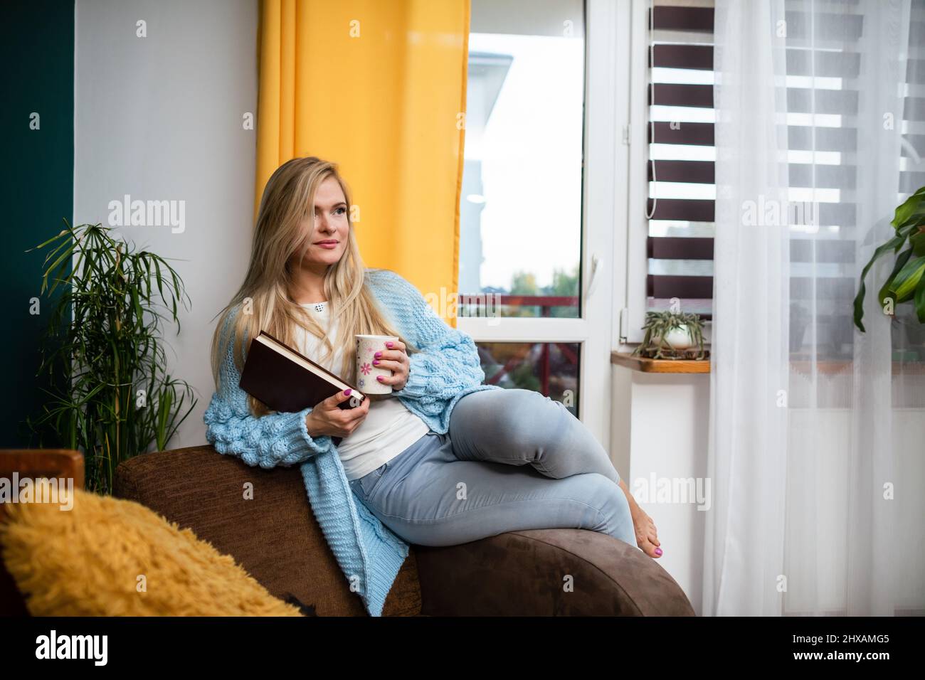 A young woman sat on the back of a chair with a book and a mug of coffee. Stock Photo