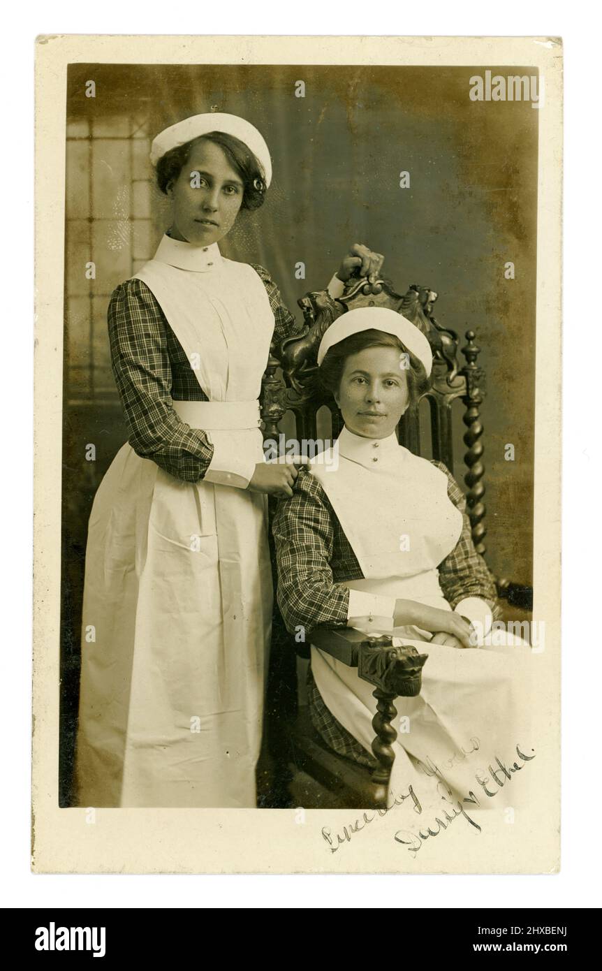 Original WW1 era postcard studio portrait of 2 / two pretty young nurses, or nursemaids in starched aprons with high bibs, caps, and detachable sleeve cuffs and collars. They both are wearing matching checked blouses, so are staff working in the same nursing home, hospital or large private house. The girls have signed Daisy and Ethel on the front, London area, U.K. circa 1915. Stock Photo