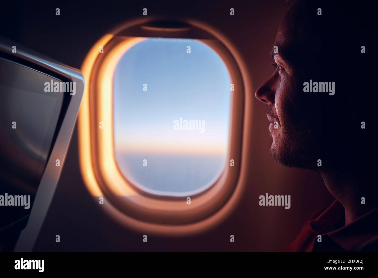 Portrait of man traveling by airplane. Passenger looking through plane window during flight at sunrise. Stock Photo