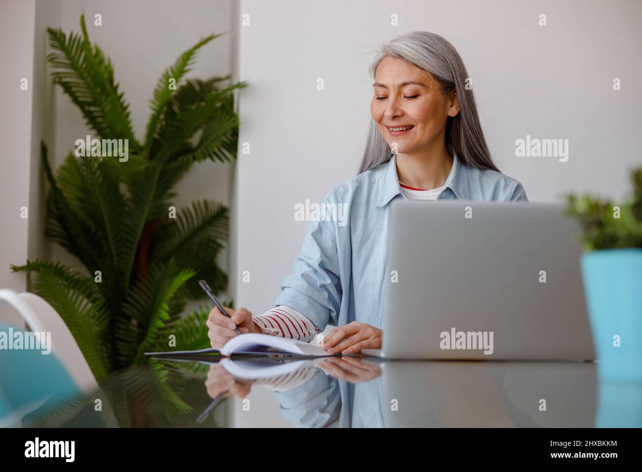 Cheerful woman writing in notebook and using laptop at home Stock Photo