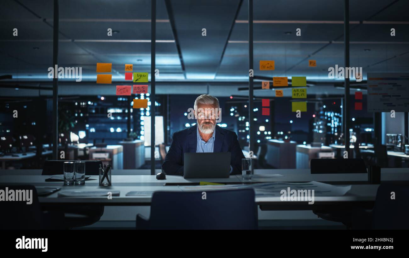 Successful Senior Businessman Working on Laptop Computer in Big City Office Late in the Evening. Investment Analyst Checking Financial Graphs from Project Management Reports. Stock Photo