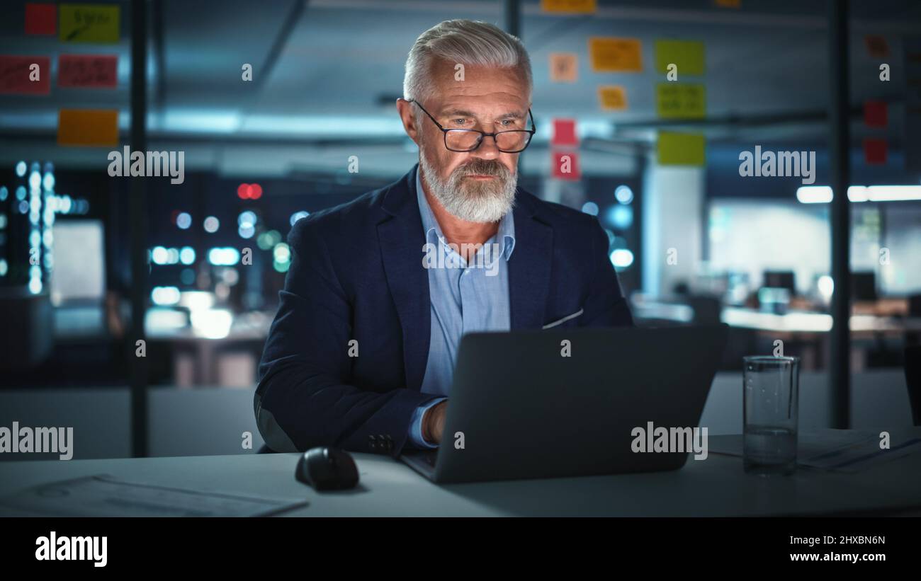 Successful Senior Businessman Working on Laptop Computer in Big City Office Late in the Evening. Investment Analyst Checking Financial Graphs from Project Management Reports. Stock Photo