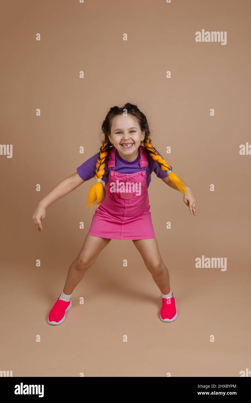 Exuberant little female child with yellow kanekalon braids with legs wide apart smiling showing piece signs with hands dressed in pink jumpsuit Stock Photo