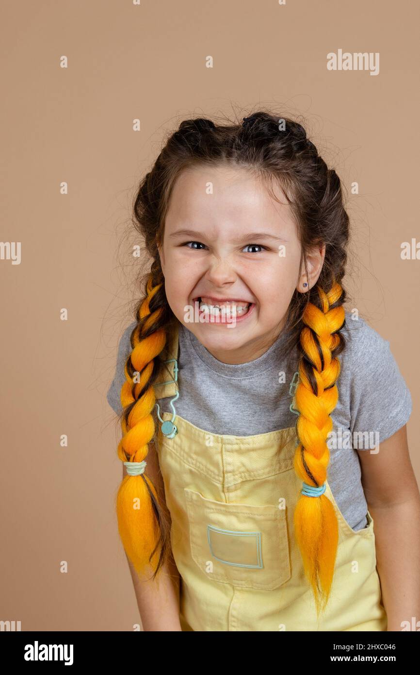 Closeup of playful indulging, little girl with yellow kanekalon pigtails, scaring someone smiling with missing tooth wearing yellow jumpsuit and gray Stock Photo