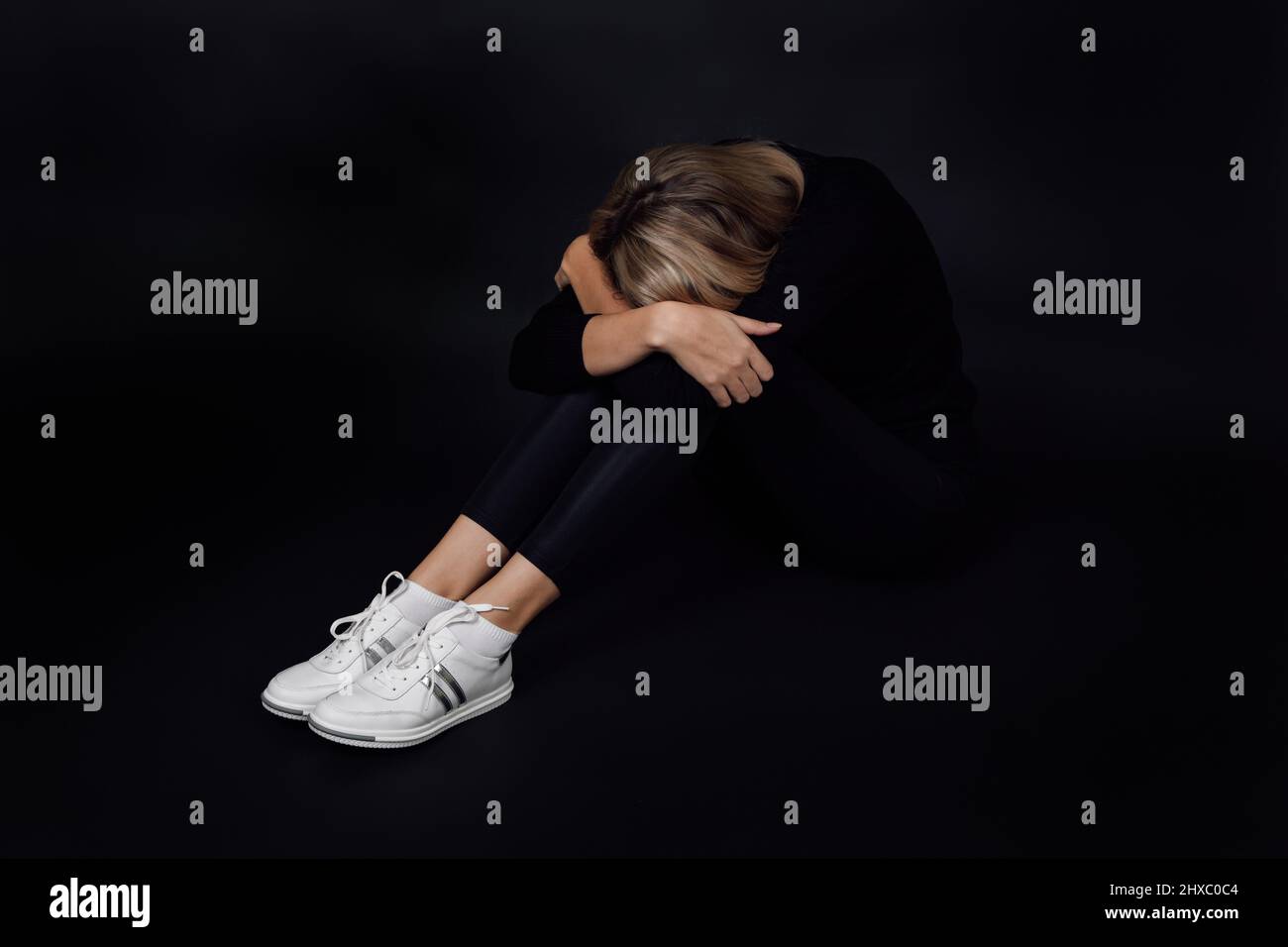 Young blonde woman in black dress going through depression with face on knees sitting on floor hiding from people on black background. Physical and Stock Photo