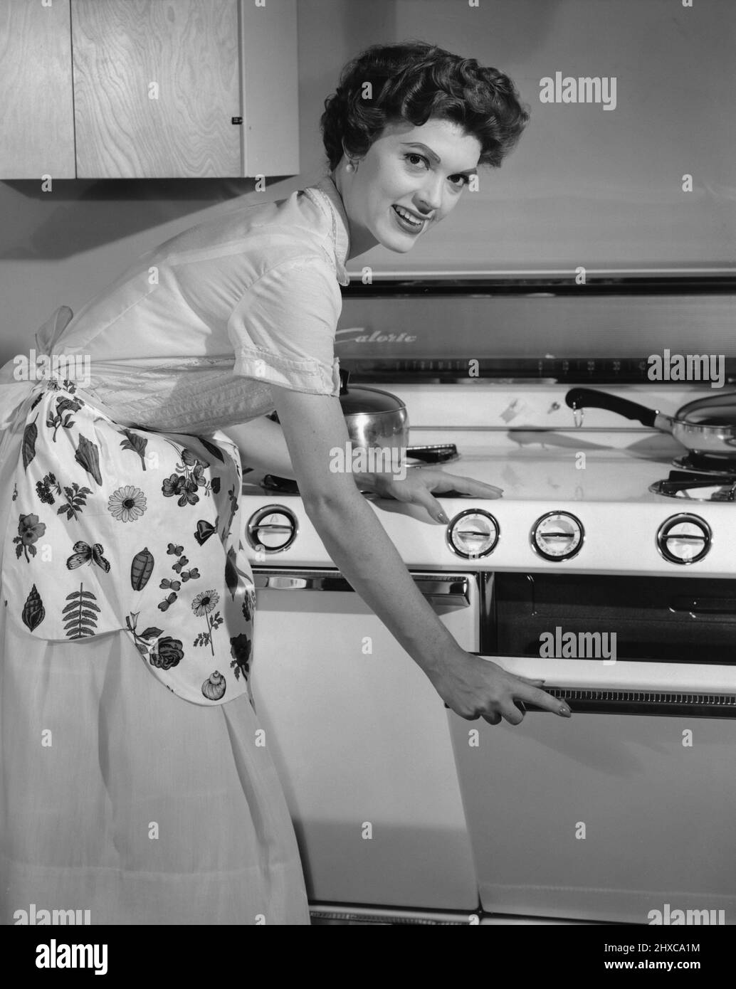 Mid-30's aged woman in a dress and apron, smiling while bending over to open oven door Stock Photo