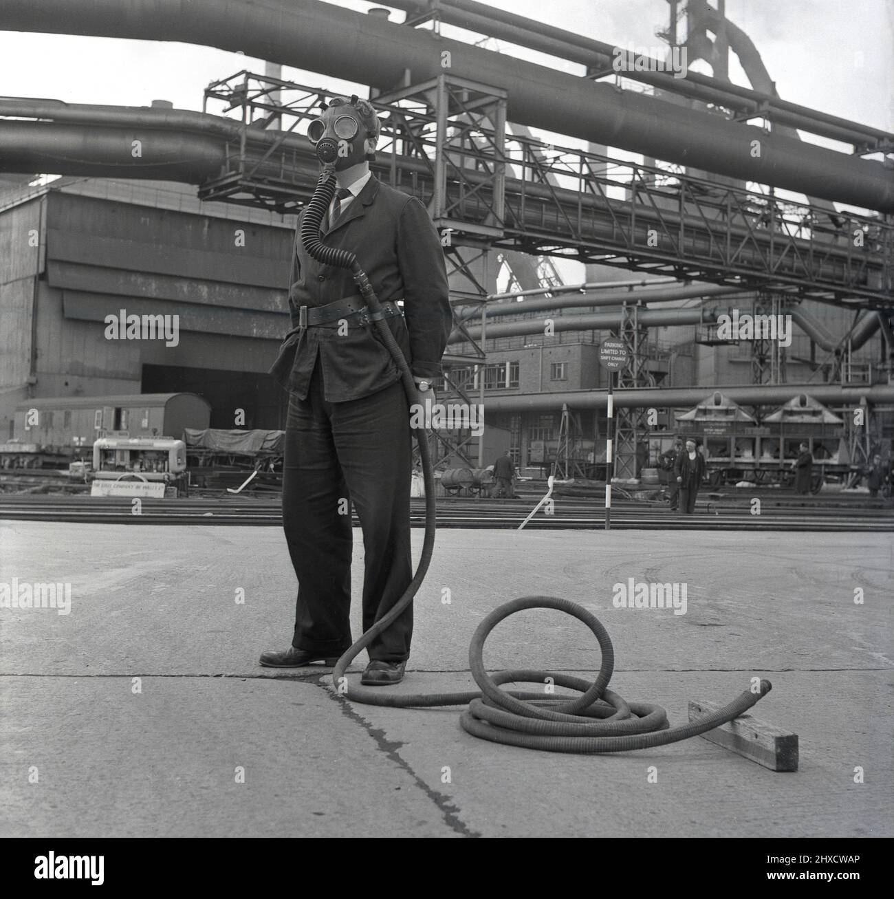 1950s, historical, at the giant Abbey Works steel complex, a worker on the tarmac outside the plant, wearing a gas mask attached to a long pipe, Port Talbot, Wales, UK. He is demonstrating the health & safety equipment available on the site from airborne pollutants and toxic gases. Stock Photo
